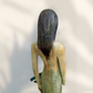 A Loretta Bertoli statue of a faceless woman in a long flowing dress with two hummingbirds, displayed against a stacked stone tile wall on a light tiled floor. This is her back view showing her hair.
