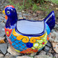 A colorful ceramic chicken-shaped planter with a glossy hand-painted finish, placed outdoors on a bed of small gray rocks and greenery, right side view.