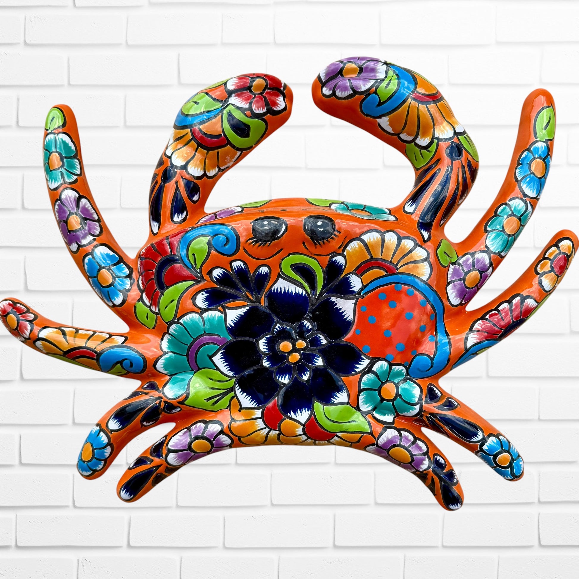 A colorful orange talavera crab wall art sculpture decorated with vibrant floral patterns, shown against a white brick wall background, top shell view.