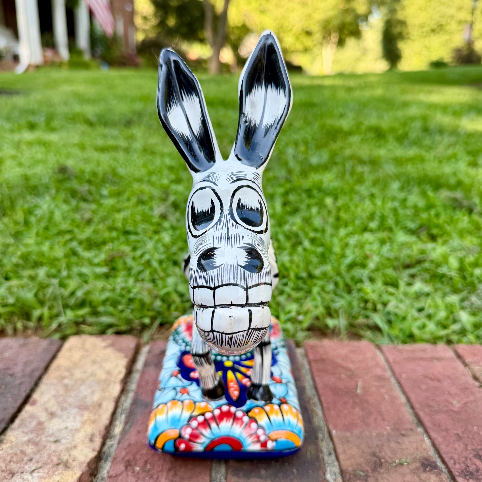 A colorful Catrina donkey figurine standing on a decorated base, photographed outdoors on a brick surface with a grassy yard and house in the background, front view of face.