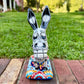 A colorful Catrina donkey figurine standing on a decorated base, photographed outdoors on a brick surface with a grassy yard and house in the background, front view of face.