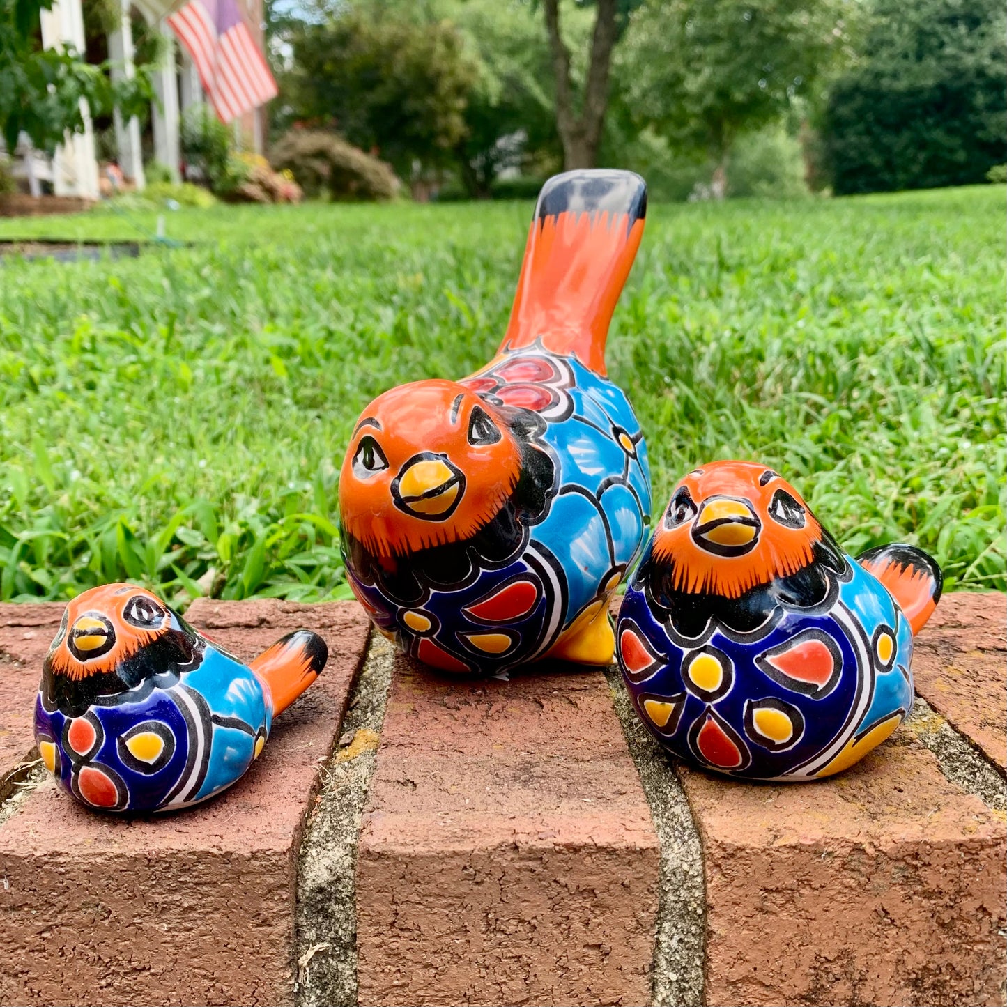 Three hand-painted ceramic bird garden statues in orange, blue, yellow, and black, displayed on a brick ledge in a backyard with a green lawn and trees in the background.