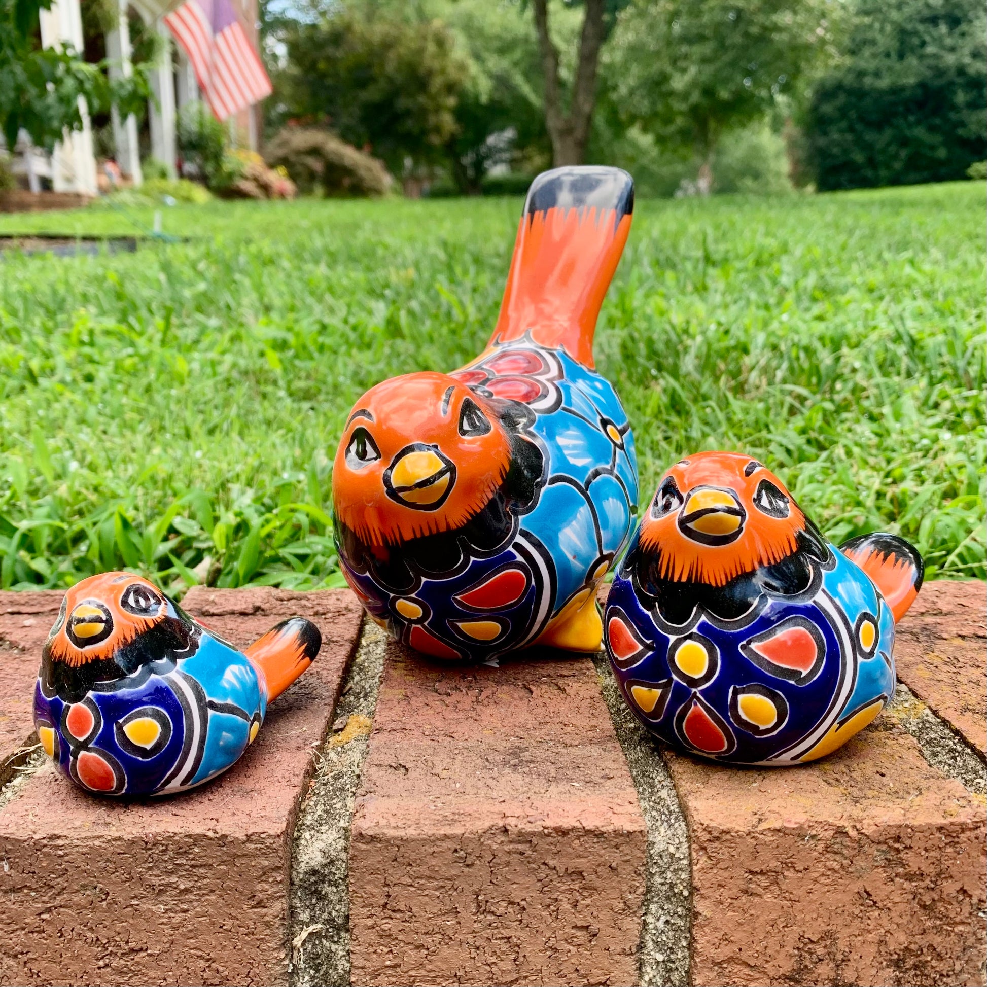 Three hand-painted ceramic bird garden statues in orange, blue, yellow, and black, displayed on a brick ledge in a backyard with a green lawn and trees in the background.
