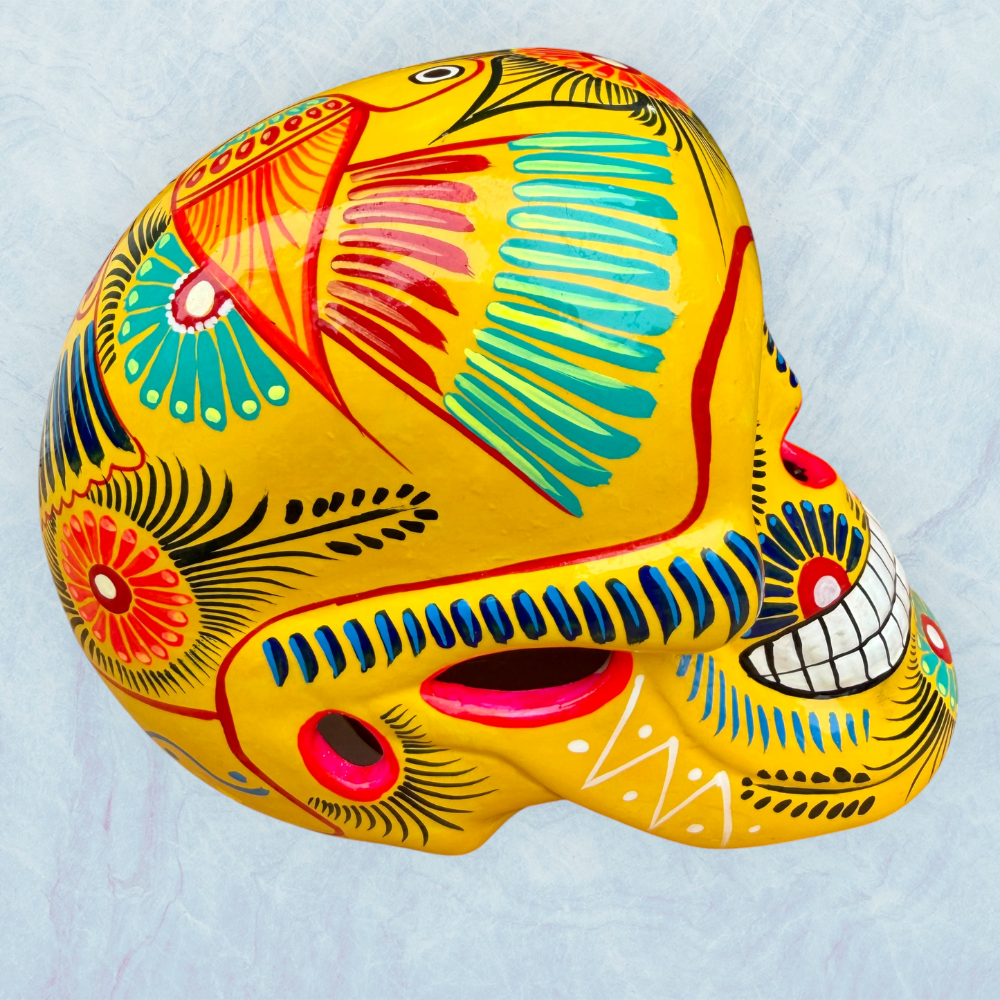 A vibrant yellow sugar skull decorated with colorful floral and geometric patterns in blue, red, black, and white, set against a light blue textured background, left side view of skull.