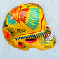 A vibrant yellow sugar skull decorated with colorful floral and geometric patterns in blue, red, black, and white, set against a light blue textured background, left side view of skull.
