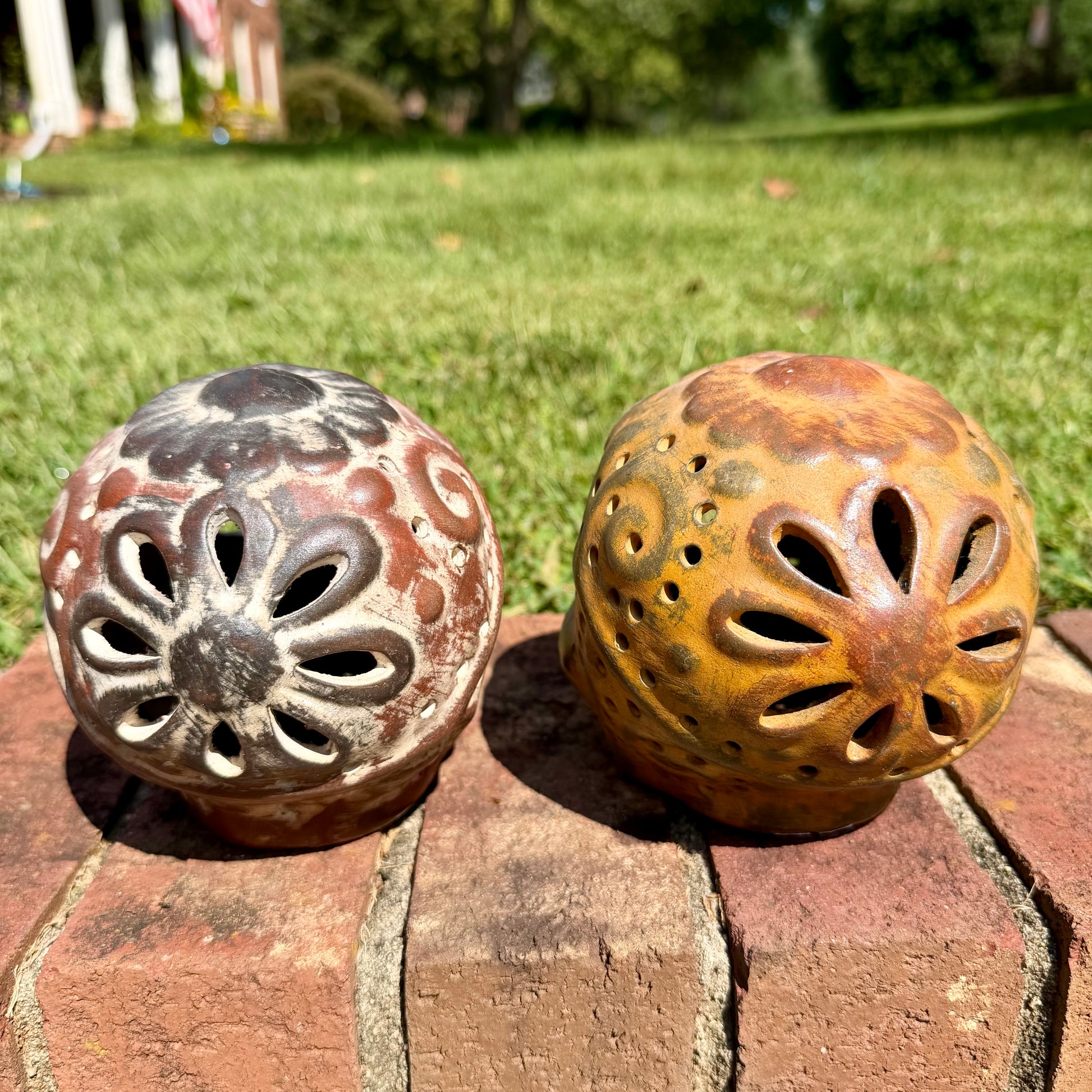 Two decorative clay sugar skull ornaments with Day of the Dead style painted patterns, displayed side-by-side on a brick surface outdoors with a grassy lawn and trees in the background, back of skull view.