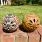 Two decorative clay sugar skull ornaments with Day of the Dead style painted patterns, displayed side-by-side on a brick surface outdoors with a grassy lawn and trees in the background, back of skull view.