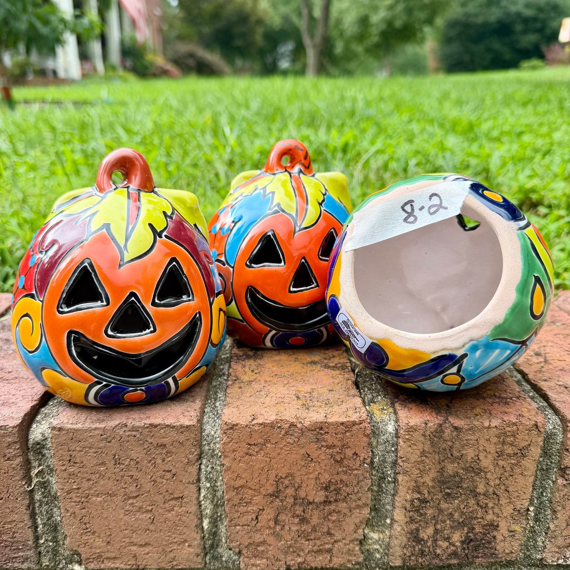 Three hand-painted Talavera ceramic jack-o'-lantern pumpkins with orange faces and yellow, blue, and red green designs, sitting on a brick path in a grassy backyard, View of bottom opening.