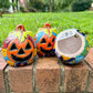 Three hand-painted Talavera ceramic jack-o'-lantern pumpkins with orange faces and yellow, blue, and red green designs, sitting on a brick path in a grassy backyard, View of bottom opening.