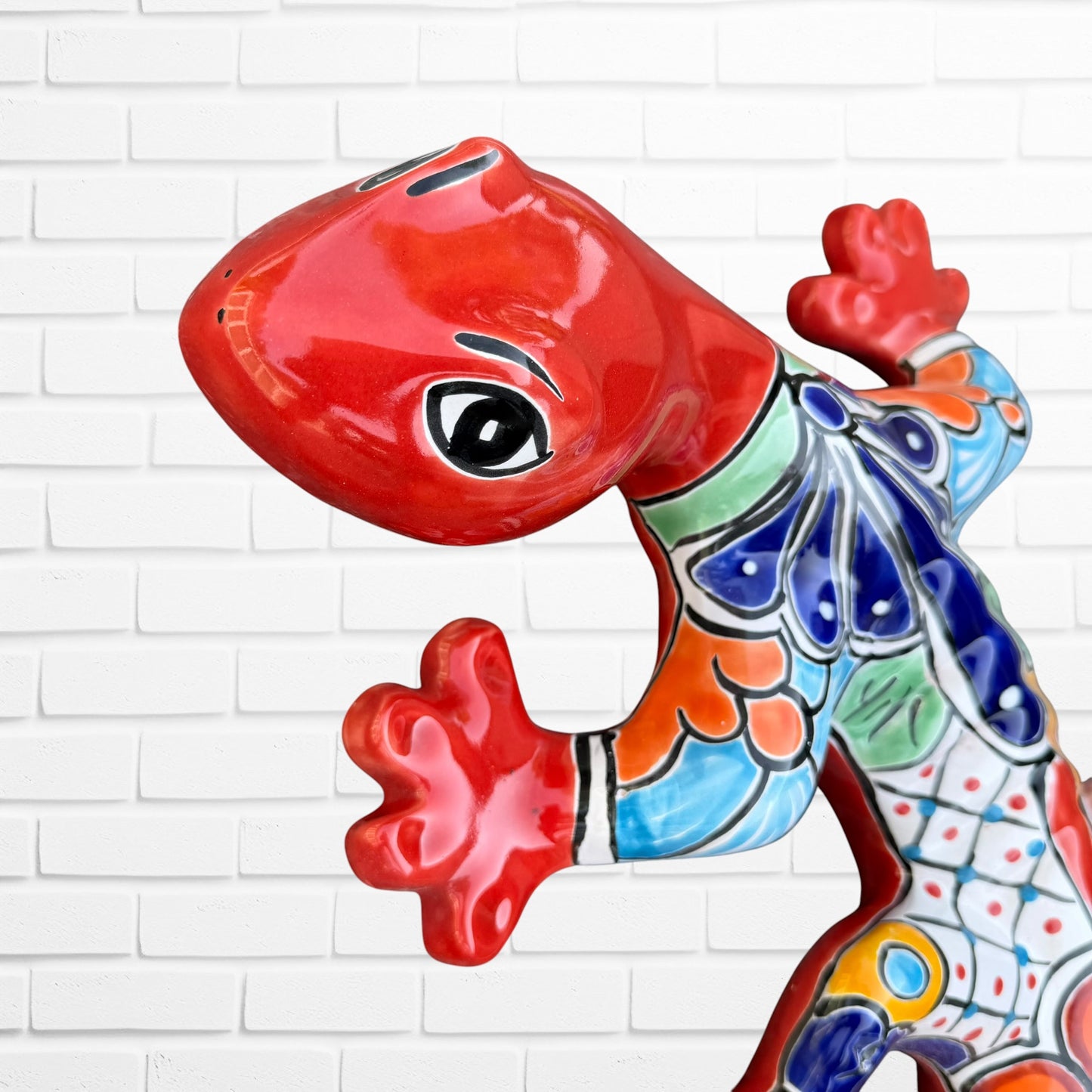 A colorful Talavera ceramic wall salamander lizard art piece with a glossy finish, featuring a red head and tail with a patterned body, displayed against a white brick wall background, close-up of face and eyes.