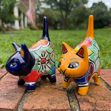Two colorful talavera ceramic cat figurines standing on a brick surface outdoors with grass and trees blurred in the background. One cat is dark blue with orange, yellow, and red patterns, and the other is orange with blue, red, yellow, and white patterns. Right side view.