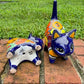 Two colorful hand-painted talavera ceramic cat figurines in cobalt blue with multi color designs standing on a brick path with a green lawn bottom of figurine view.