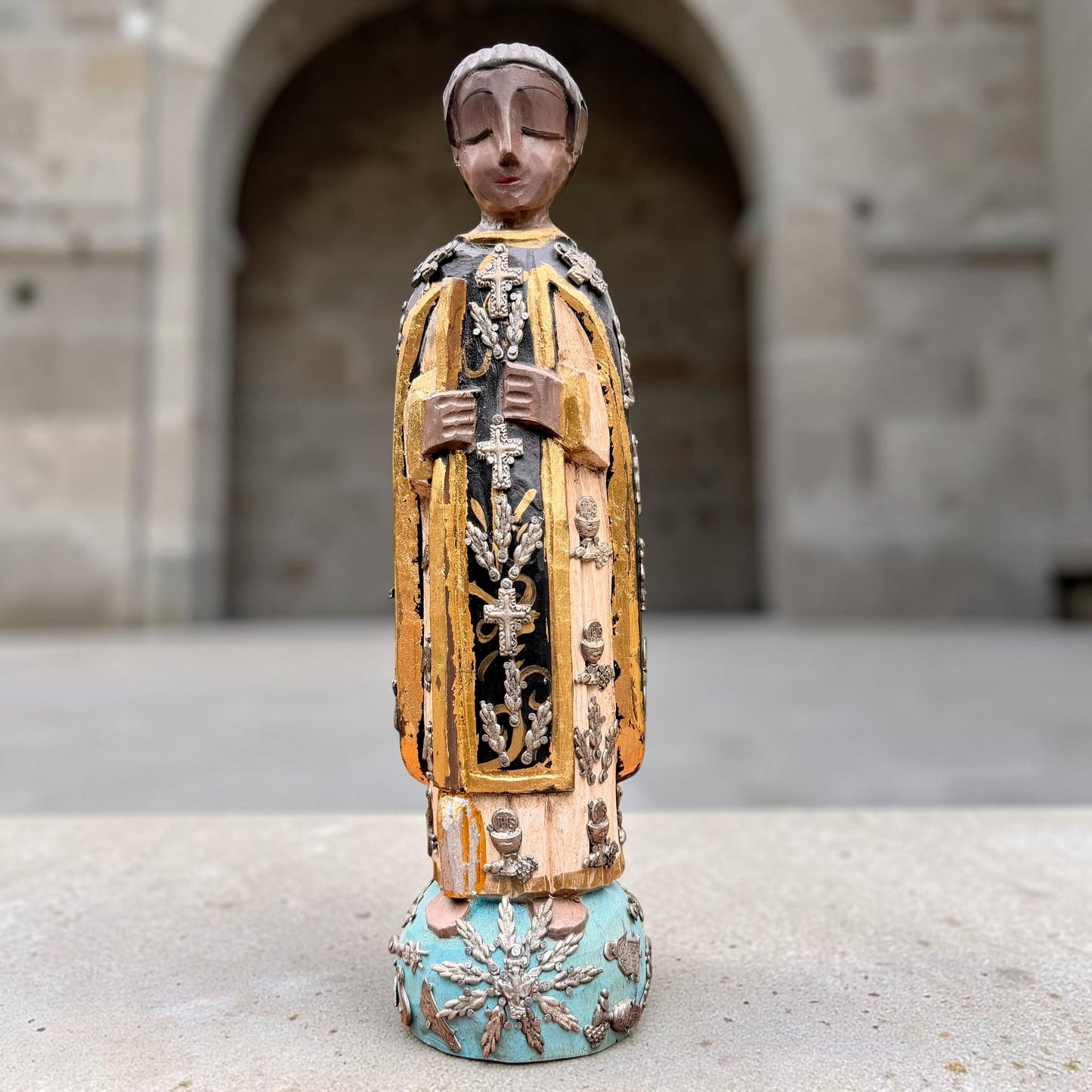 A carved milagros religious figurine of  Saint Martin in ornate black and gold vestments with silver cross details, photographed outdoors in front of a blurred stone archway. Full front view of figurine.