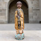A carved milagros religious figurine of  Saint Martin in ornate black and gold vestments with silver cross details, photographed outdoors in front of a blurred stone archway. Full front view of figurine.