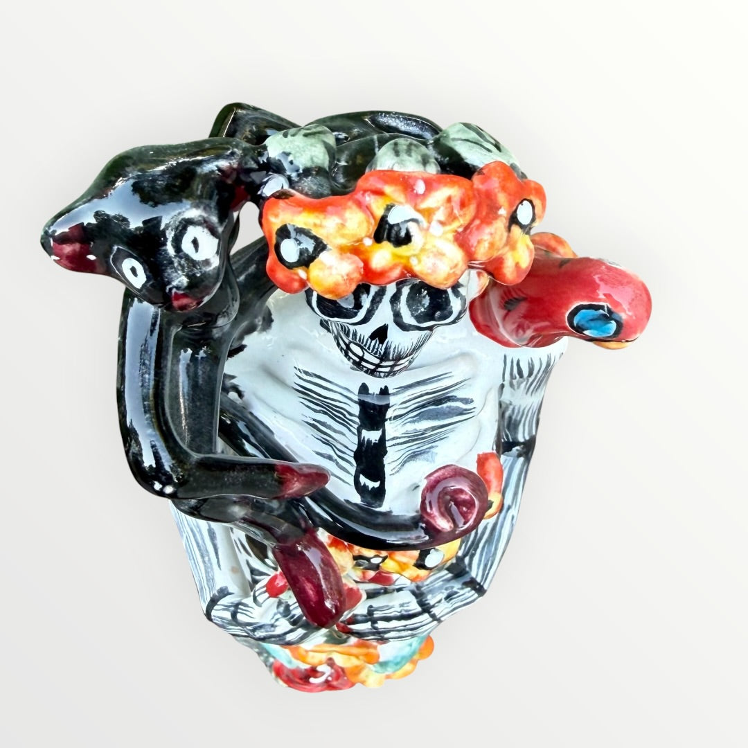 A Day of the Dead Catrina of a Frida skeleton doll holding a black cat, with a red parrot on her shoulder and colorful flowers around the top of her dress. Top view of Frida showing monkey, parrot and floral headband.