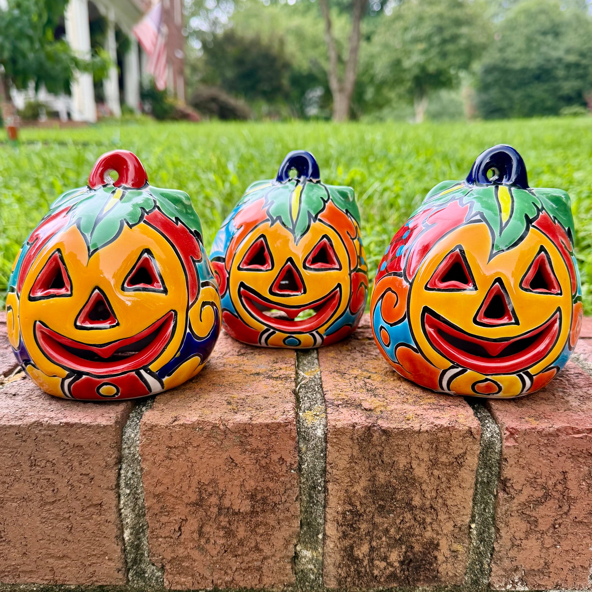 Three hand-painted Talavera ceramic jack-o'-lantern pumpkins in bright orange, yellow, red, green, and blue, sitting on a brick ledge with a blurred green lawn and house in the background.
