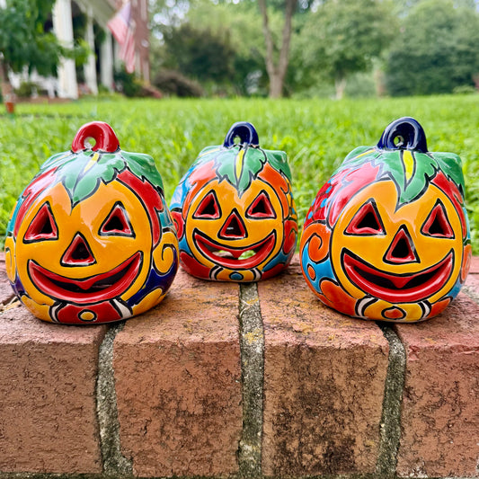 Three hand-painted Talavera ceramic jack-o'-lantern pumpkins in bright orange, yellow, red, green, and blue, sitting on a brick ledge with a blurred green lawn and house in the background.