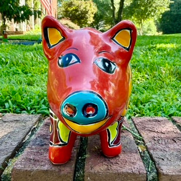 A colorful hand-painted red Talavera ceramic pig figurine standing on a brick surface with a blurred green lawn and trees in the background face close up