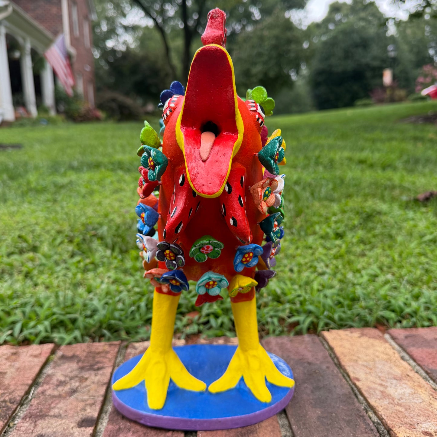 A colorful decorative rooster figurine standing on a brick surface outdoors, front view with open beak, with a grassy lawn and trees in the background. The rooster is primarily red and yellow with many small multicolored flowers and painted details across its body and tail.