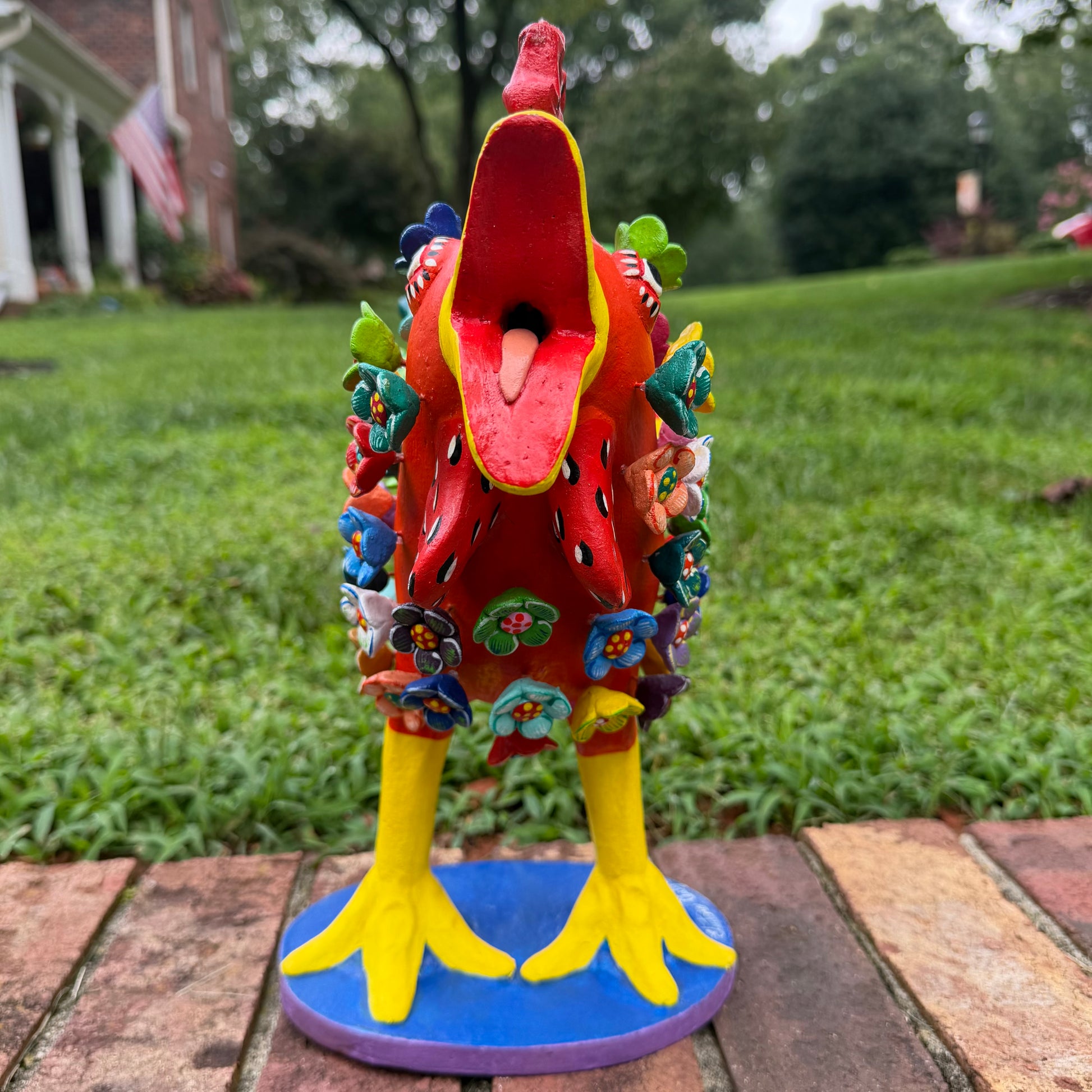 A colorful decorative rooster figurine standing on a brick surface outdoors, front view with open beak, with a grassy lawn and trees in the background. The rooster is primarily red and yellow with many small multicolored flowers and painted details across its body and tail.