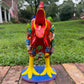 A colorful decorative rooster figurine standing on a brick surface outdoors, front view with open beak, with a grassy lawn and trees in the background. The rooster is primarily red and yellow with many small multicolored flowers and painted details across its body and tail.