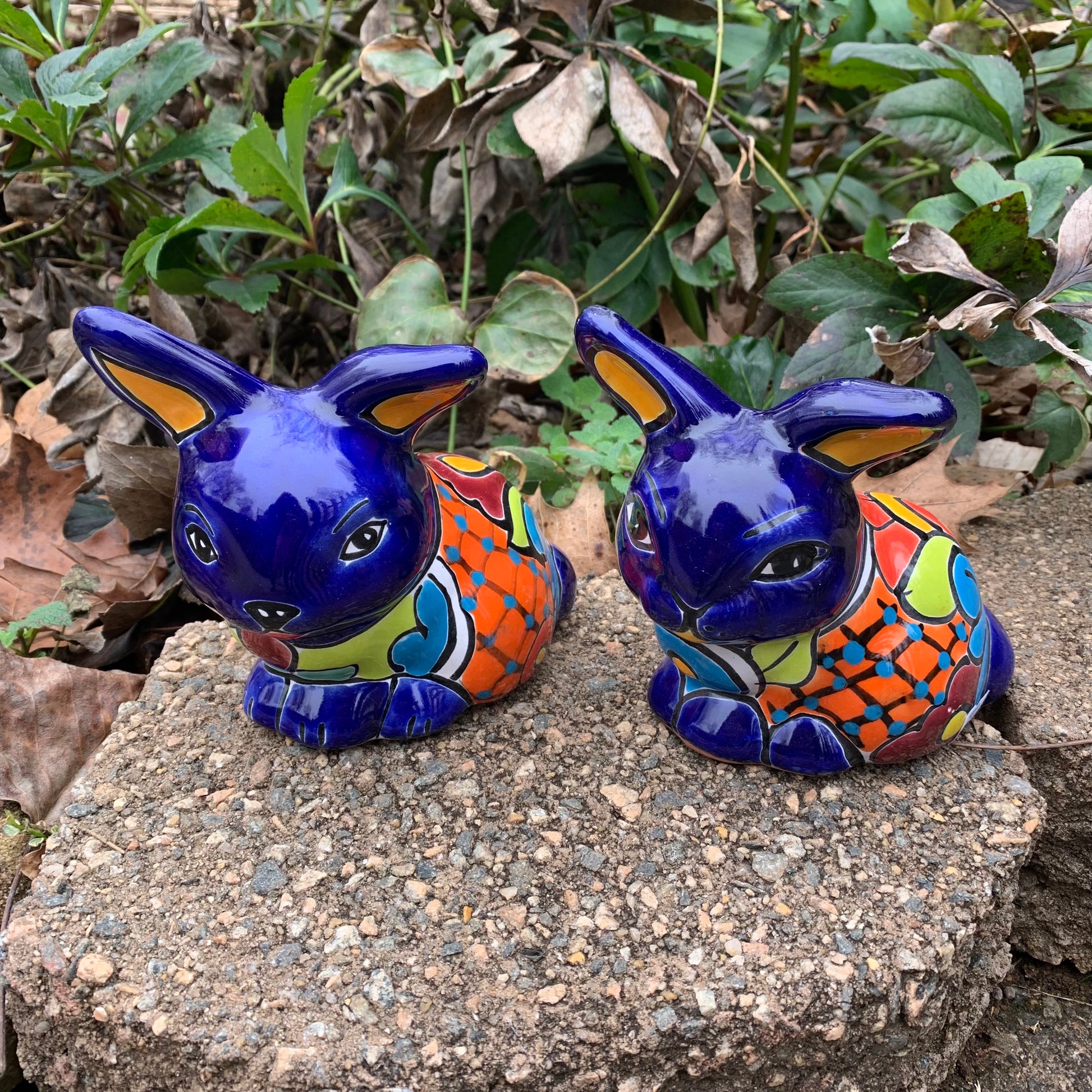 Two colorful Mexican Talavera rabbit figurines on a stone surface with greenery in the background set of two azul