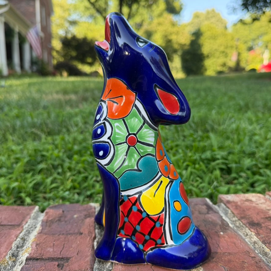 A colorful hand-painted Mexican Talavera Ceramic dog figurine sitting on a brick wall with a green grassy lawn and a house in the background side view