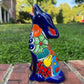 A colorful hand-painted Mexican Talavera Ceramic dog figurine sitting on a brick wall with a green grassy lawn and a house in the background side view
