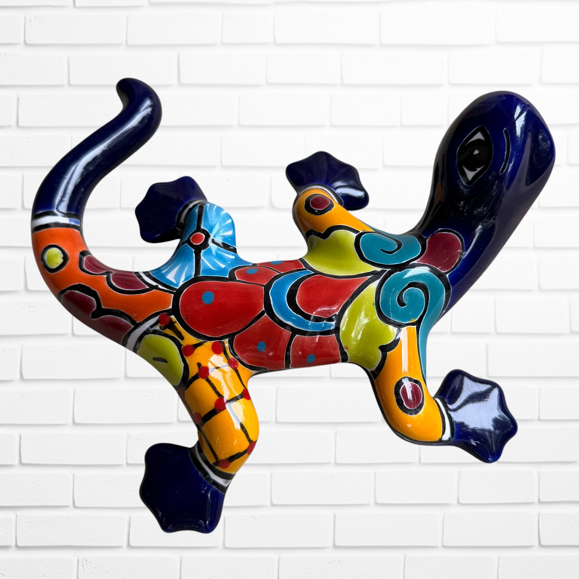 A colorful ceramic wall sculpture of a stylized lizard or salamander, decorated with vibrant hand-painted patterns in blue, red, yellow, and orange, mounted on a white brick wall back side.