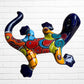 A colorful ceramic wall sculpture of a stylized lizard or salamander, decorated with vibrant hand-painted patterns in blue, red, yellow, and orange, mounted on a white brick wall back side.