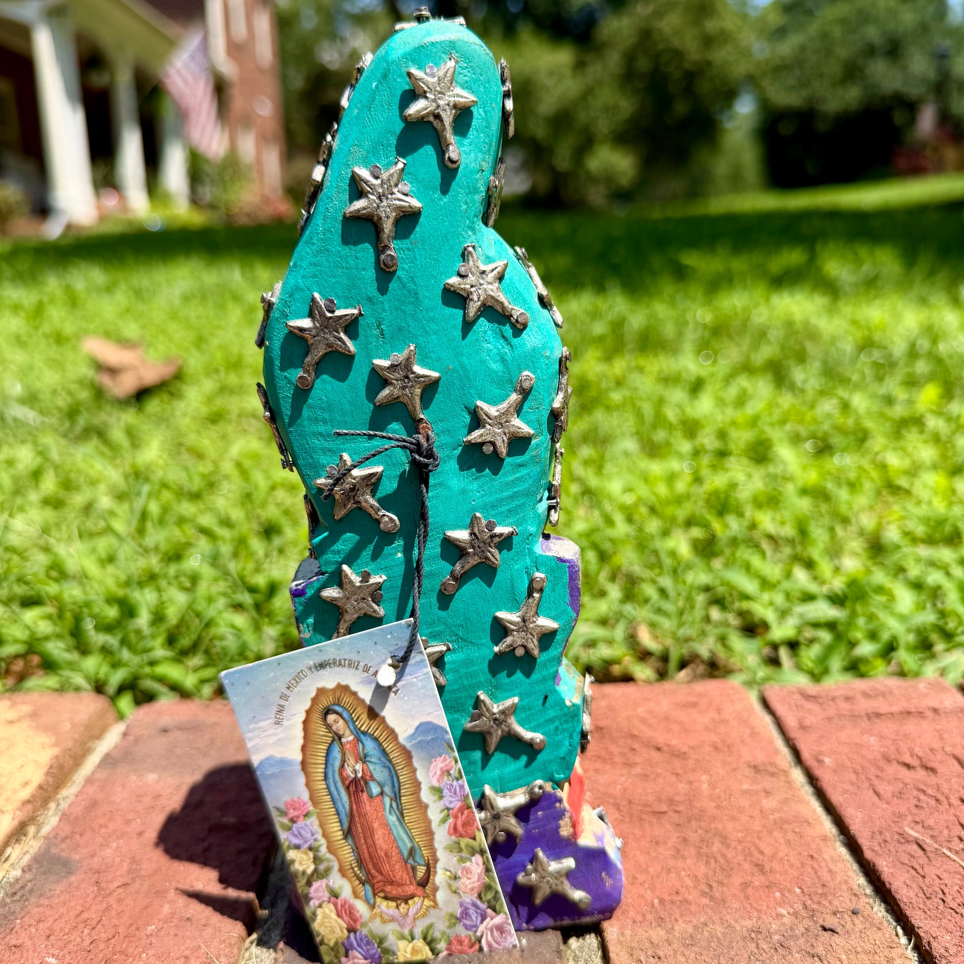 A colorful Milagros Guadalupe  (Virgin Mary) standing on a brick surface outdoors with a grassy yard in the background, back view of Guadalupe.