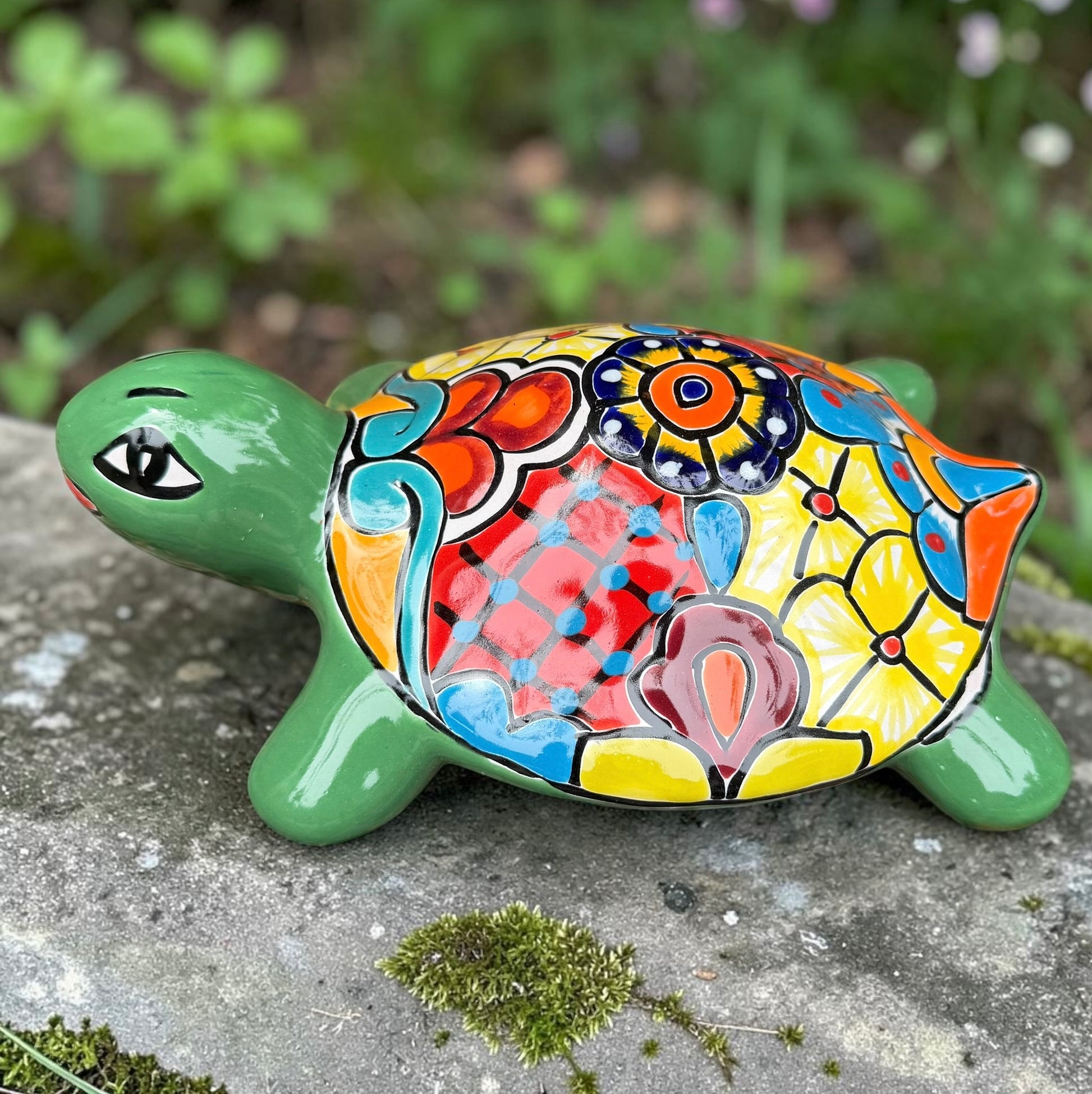 A hand-painted ceramic turtle figurine resting on a gray stone surface with a blurred green garden background. The turtle features a bright green head and legs, and a highly detailed, multicolored shell with yellow, red, orange, blue, and black patterns, right side view.