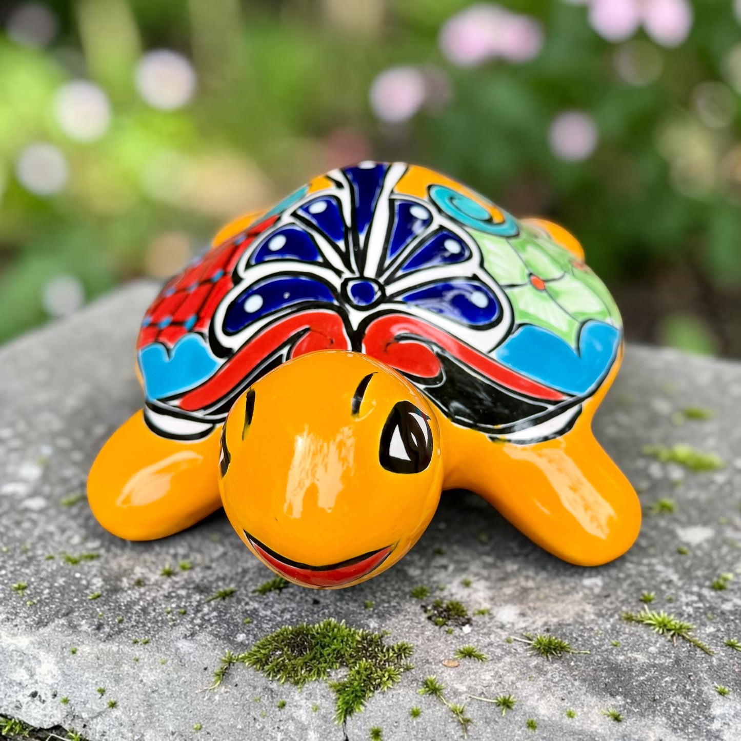 A glossy ceramic turtle figurine with a bright yellow body and a highly decorated shell in red, blue, yellow, black, and white, resting on a stone surface with a blurred garden background, front view.