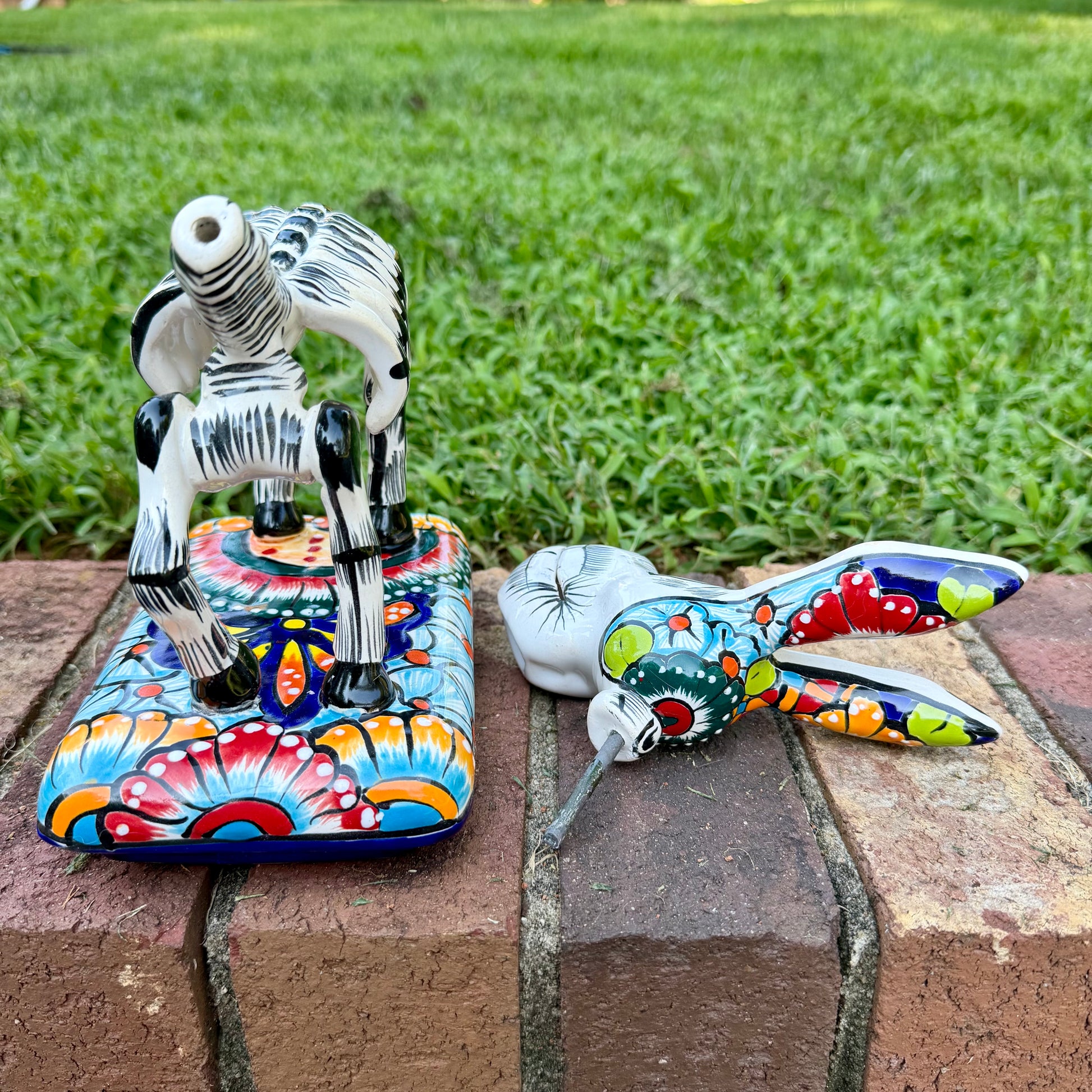 A colorful Catrina donkey figurine standing on a decorated base, photographed outdoors on a brick surface with a grassy yard and house in the background, Photo of donkey base with head removed.