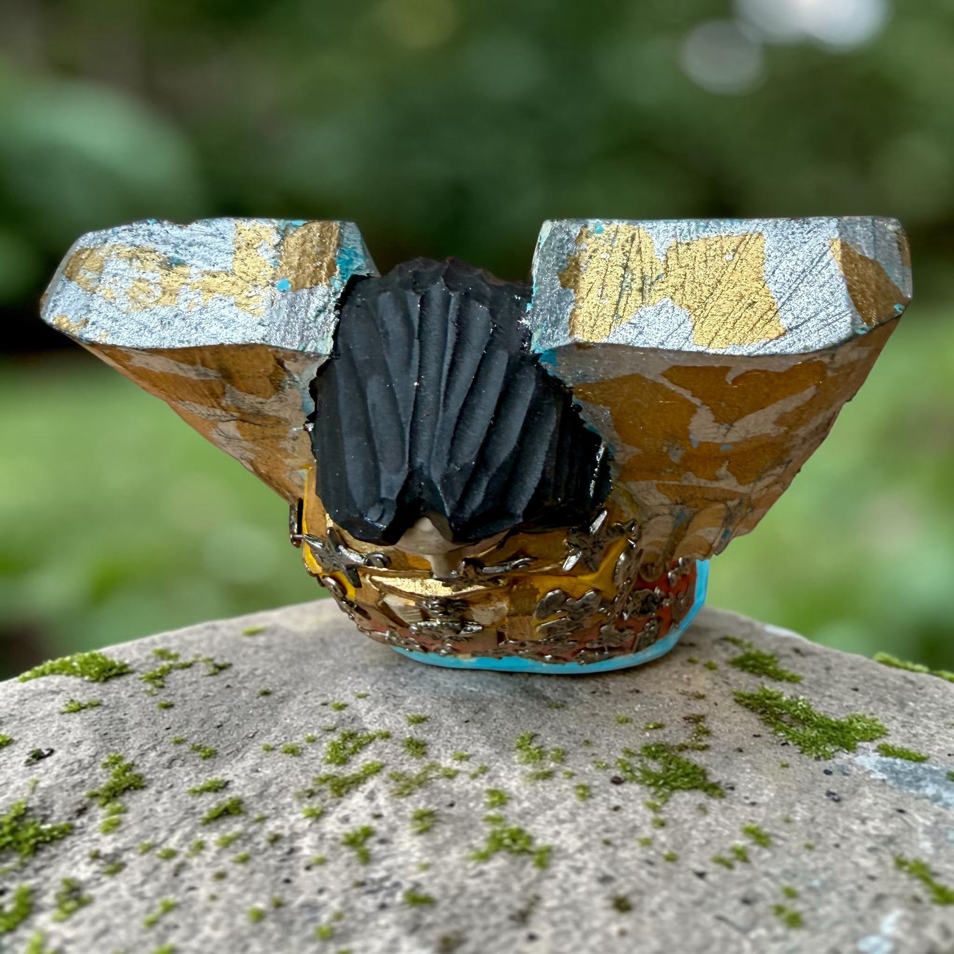 A handcrafted wooden Milagros angel figurine with large wings, decorated with metallic silver charms and painted in yellow, and orange, standing on a mossy stone with a blurred green garden background. Top view of angel from wings.