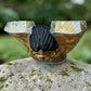 A handcrafted wooden Milagros angel figurine with large wings, decorated with metallic silver charms and painted in yellow, and orange, standing on a mossy stone with a blurred green garden background. Top view of angel from wings.