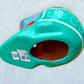 A decorative skull sculpture with intricate colorful sugar skull style patterns painted on a teal colored base, set against a light blue textured background, bottom view: