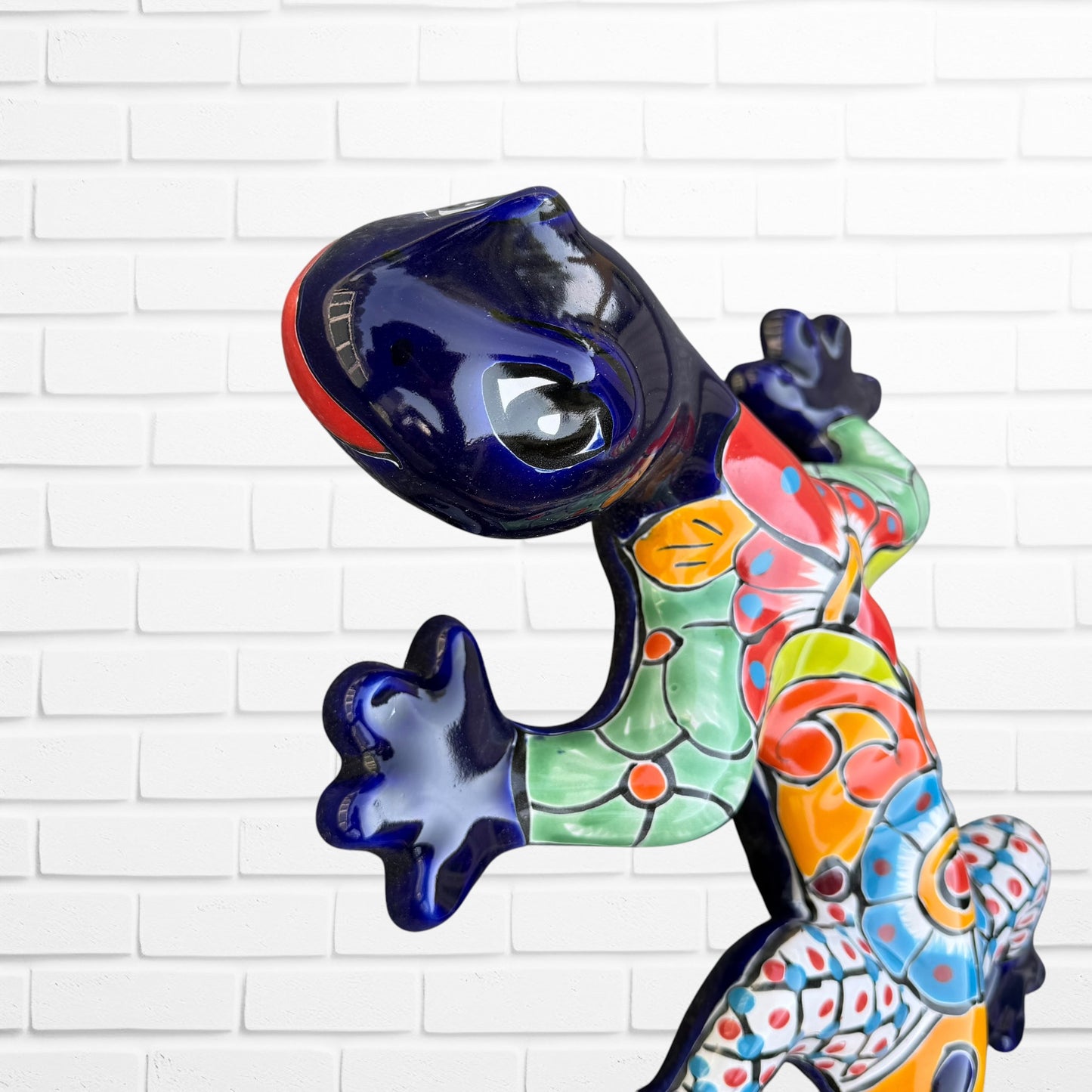 A colorful Mexican Talavera wall sculpture of a lizard, featuring a vibrant multicolor pattern with dark blue, orange, red, green, and yellow, mounted on a white brick wall, close-up of face and eyes.