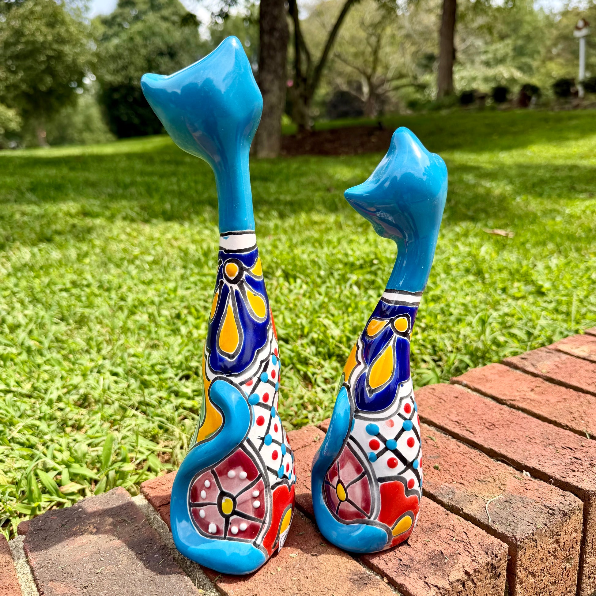 Two colorful ceramic cat figurines standing on a brick path in a grassy outdoor garden setting. This is their back view, including tails.