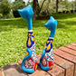 Two colorful ceramic cat figurines standing on a brick path in a grassy outdoor garden setting. This is their back view, including tails.