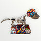 A ceramic Mexican-style skeleton dog figurine standing on a colorful floral base against a plain white background. Left side view of catrina skeleton dog.