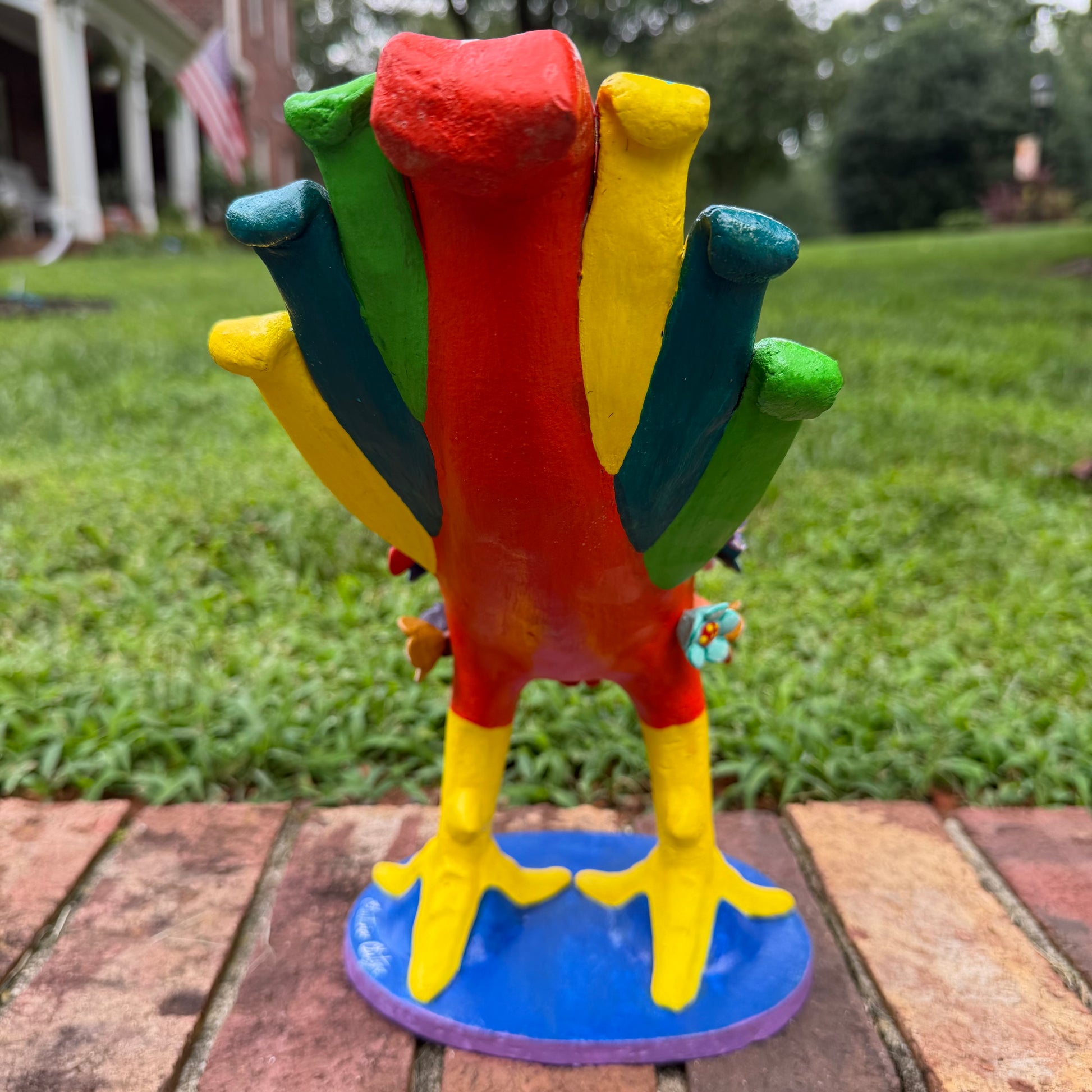 A colorful decorative rooster figurine standing on a brick surface outdoors, back tail view, with a grassy lawn and trees in the background. The rooster is primarily red and yellow with many small multicolored flowers and painted details across its body and tail.