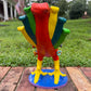 A colorful decorative rooster figurine standing on a brick surface outdoors, back tail view, with a grassy lawn and trees in the background. The rooster is primarily red and yellow with many small multicolored flowers and painted details across its body and tail.