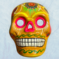 A clay, yellow with floral designs, Mexican-style skeleton skull face mask hanging on a soft blue surface.