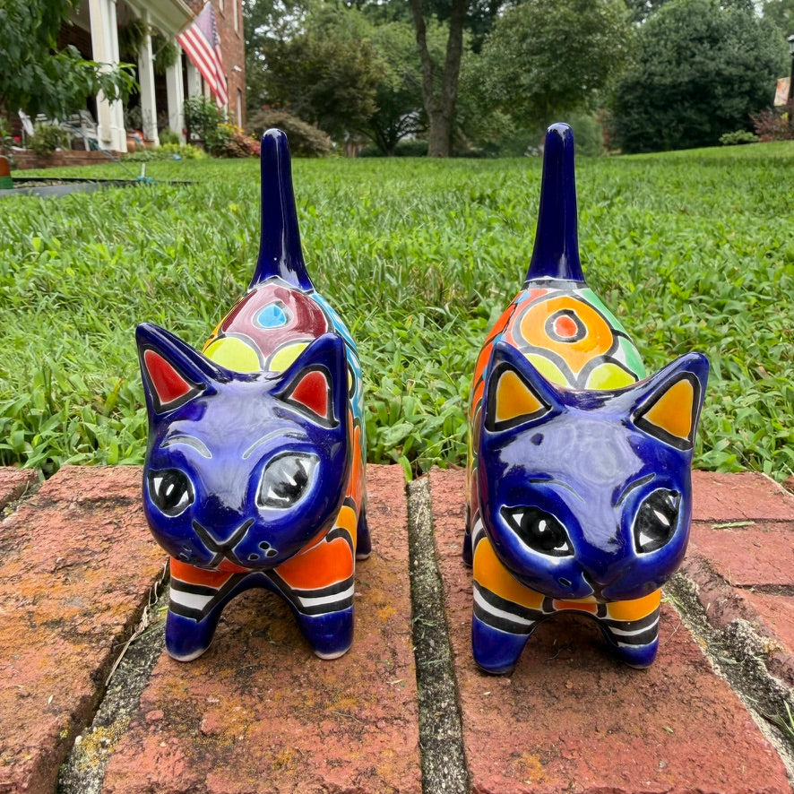 Two colorful hand-painted talavera ceramic cat figurines in cobalt blue with multi color designs standing on a brick path with a green lawn front face view.