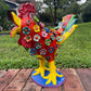 A colorful decorative rooster figurine standing on a brick surface outdoors, with a grassy lawn and trees in the background. The rooster is primarily red and yellow with many small multicolored flowers and painted details across its body and tail.