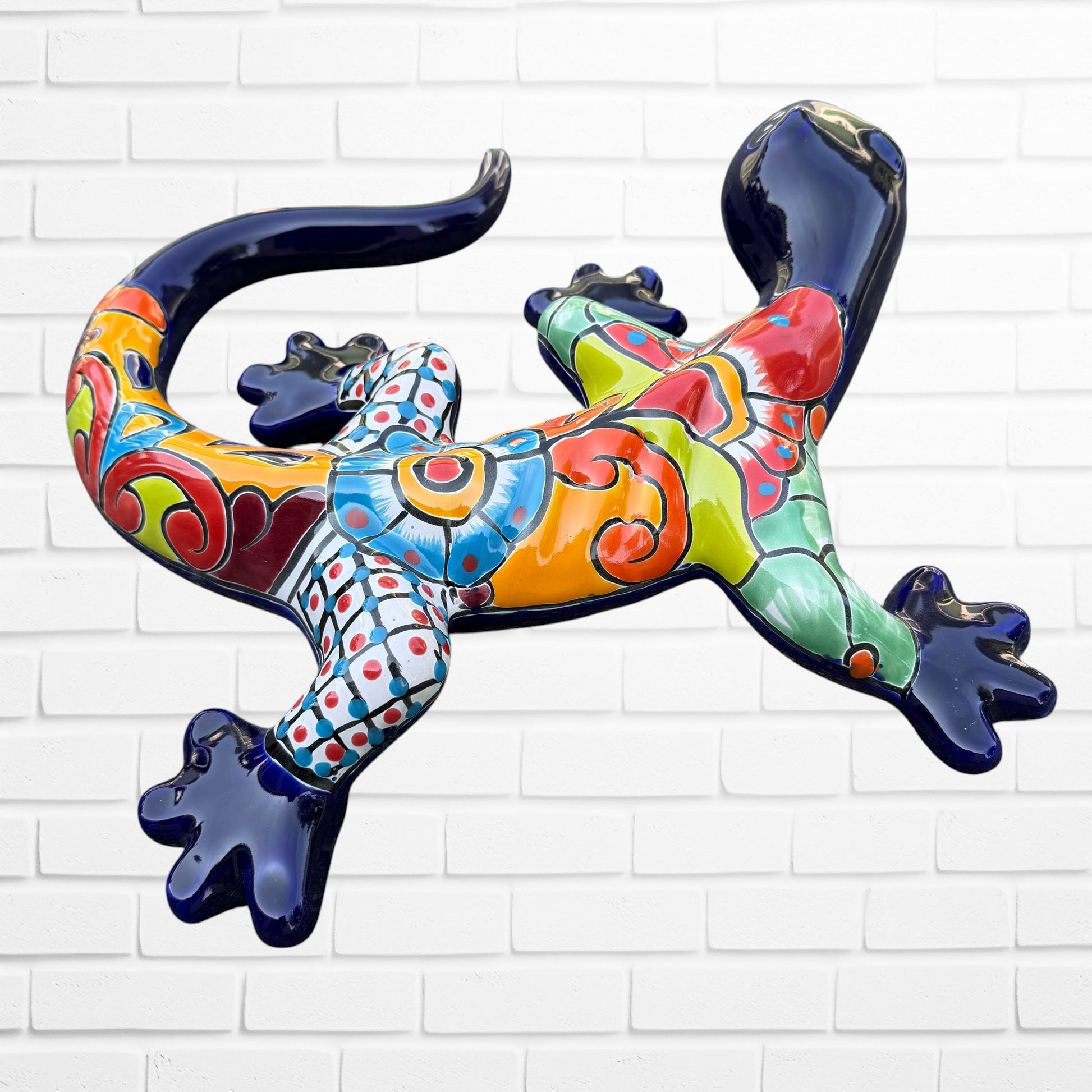 A colorful Mexican Talavera wall sculpture of a lizard, featuring a vibrant multicolor pattern with dark blue, orange, red, green, and yellow, mounted on a white brick wall, right side view.
