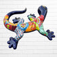 A colorful Mexican Talavera wall sculpture of a lizard, featuring a vibrant multicolor pattern with dark blue, orange, red, green, and yellow, mounted on a white brick wall, right side view.