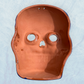 A clay, purple with designs, Mexican-style skeleton skull face mask hanging on a soft blue surface, back view.