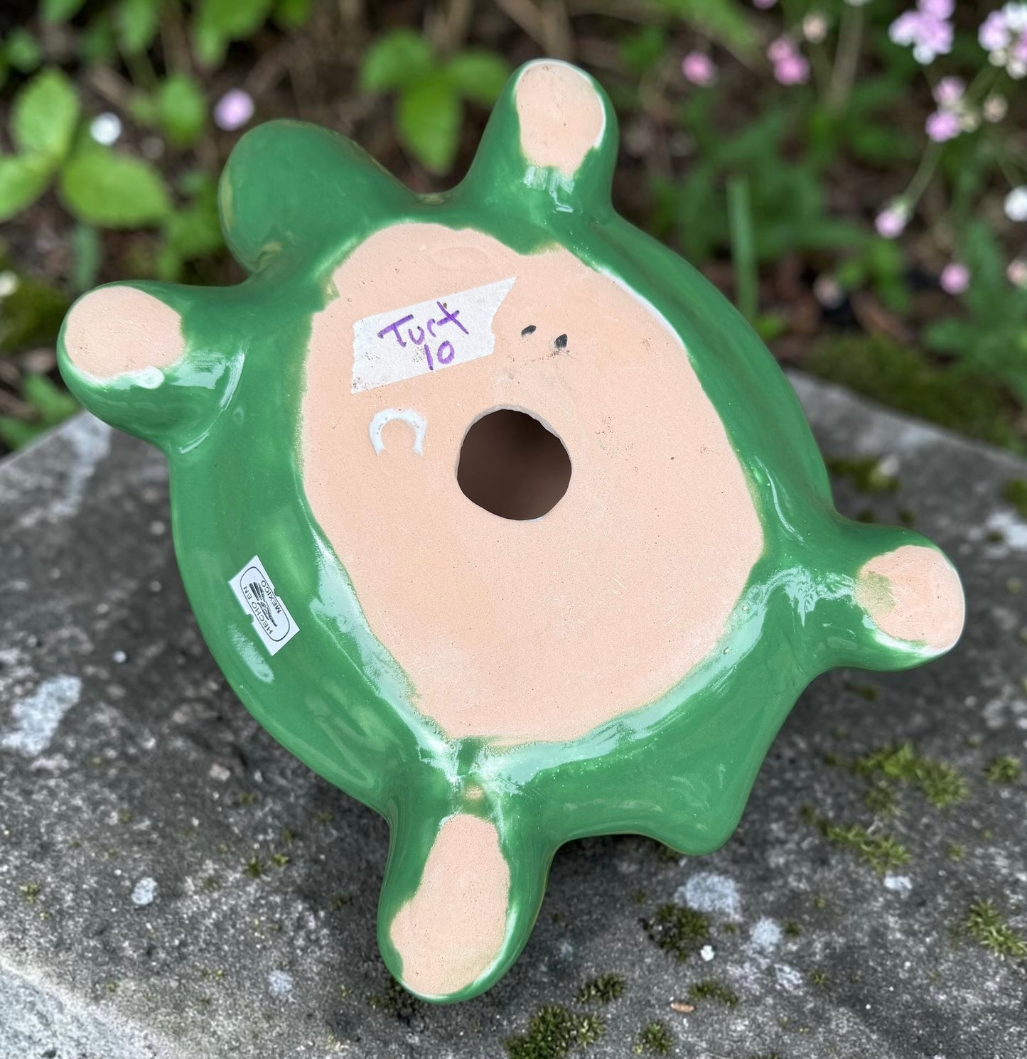 A hand-painted ceramic turtle figurine resting on a gray stone surface with a blurred green garden background. The turtle features a bright green head and legs, and a highly detailed, multicolored shell with yellow, red, orange, blue, and black patterns, bottom view of turtle,